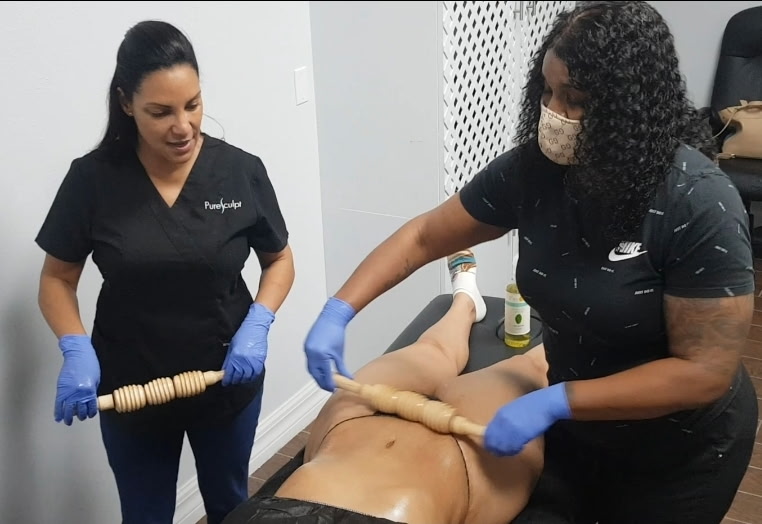 Two gloved people hold wooden massage rollers beside a client on a treatment table.